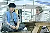 Two men sit at a wooden picnic table under a Coca-Cola umbrella in an outdoor setting.