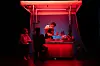 Small food stall under red light with seated customers and a vendor preparing food behind the counter.
