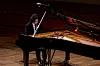 A person seated at a black grand piano on a wooden stage.