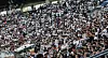 A packed baseball stadium crowd fills the stands, many wearing white shirts and cheering during a game.