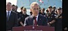A man speaks at a podium outdoors with military personnel standing in formation behind him.