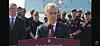 A man speaks at a podium outdoors with military personnel standing in formation behind him.