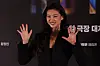 Jung Ho-yeon smiles and waves with both hands at a movie event backdrop.