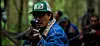 Man in a cap and denim jacket aims a rifle in a forest with blurred people behind him.