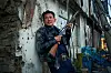 Man holding a rifle stands by a damaged wall in an urban alley.