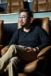 Man wearing glasses and a black shirt seated in a brown leather chair in a dim interior.