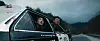 Two people lean out of a car on a mountain road under heavy storm clouds.