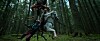 A white horse with a rider stands in a forest beside another person.