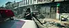 A person holds a gun beside a car on a debris-strewn street lined with damaged buildings.