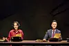 Two presenters seated behind a table on a dark stage with papers and microphones.