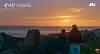 Two people view a sunrise over the sea from a rocky shoreline.