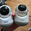A hand holds two white and black home security cameras side by side indoors.
