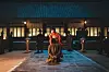 A man in traditional dress stands in a dim palace courtyard with guards and lanterns at night.