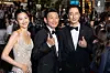 Three people in evening wear posing on a red carpet at a crowded event.