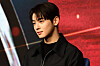 Cha Eun-woo speaking at a press event in front of a red promotional backdrop.
