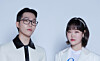 Two people in white and black outfits standing against a light studio background.
