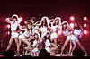 Girls' Generation performs onstage in white outfits under bright red concert lights.