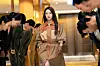 Woman in a tan trench coat walks down a hallway between two rows of people bowing toward her.