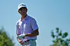 Golfer in a white cap and pink shirt stands on a golf course holding a towel during practice.