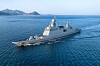 A gray naval warship moves through blue ocean waters with mountains visible in the distance.