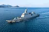 A gray naval warship moves through blue ocean waters with mountains visible in the distance.