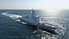 A gray naval ship sailing across the ocean with a long wake trailing behind.