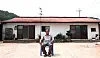 A man seated in front of a small house with a red roof and concrete yard.