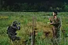 Two camouflaged soldiers stand in a grassy field near a mounted device and observe the area.