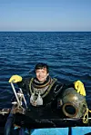 A diver in yellow gloves sits on a boat with diving equipment against a calm ocean backdrop.