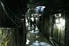Two figures walking through a wet, dimly lit tunnel with pipes and lights along the walls.