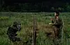 Two uniformed personnel in a field, one aiming a long gun and the other standing nearby with a piece of paper.