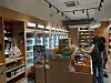 Two shoppers browse products in a convenience store with refrigerators and wooden display shelves.