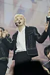 A blonde performer in a black jacket sings into a headset mic on stage.