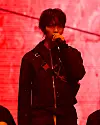 Performer in dark clothing sings into a microphone against a red illuminated backdrop.