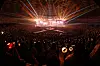 Large crowd in a dark arena facing a brightly lit concert stage with colorful laser lights.