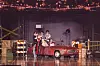 Red vintage car on a wet stage with people and props under theatrical lights.
