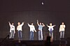 Six performers standing in a row on a dark stage, waving and raising their arms.