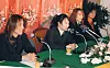 Four band members seated at microphones behind a flower-decorated table with a red backdrop.