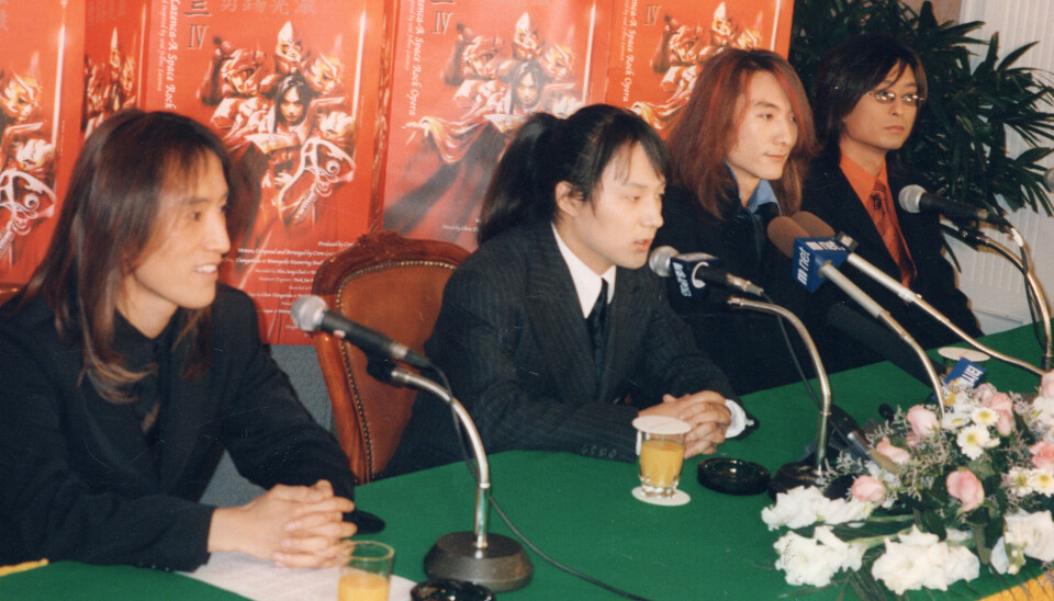 Four band members seated at microphones behind a flower-decorated table with a red backdrop.