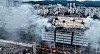 A large logistics warehouse is surrounded by thick smoke as firefighters respond from below.