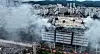 A large logistics warehouse is surrounded by thick smoke as firefighters respond from below.