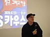 A man in a black cap stands in front of a projected event poster with Korean text.