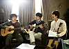 Three young men seated indoors playing acoustic guitars in a sunlit living room.
