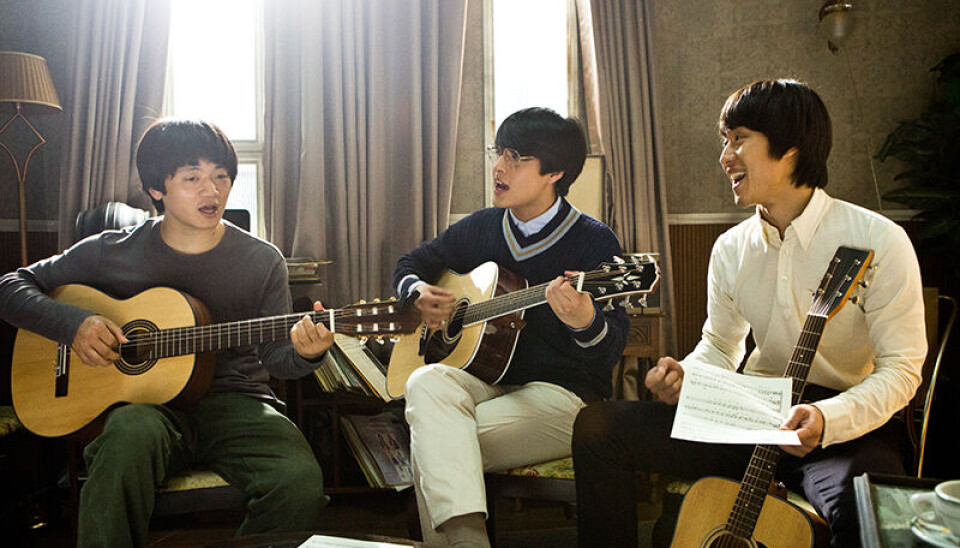 Three young men seated indoors playing acoustic guitars in a sunlit living room.