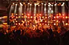 Rock band playing on stage before an audience under bright orange concert lighting.