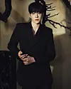 Man in a black suit standing against a beige wall with dark branch shadows.