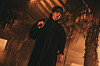 Person in a long dark coat standing in a dim industrial room with orange lighting.