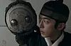 Young man looks toward a large mask with round eyes in a dark room.
