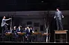 Actors in period costumes perform on a classroom-style stage during a play press call.