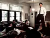 A man standing in front of a classroom speaks to students seated at desks near large windows.